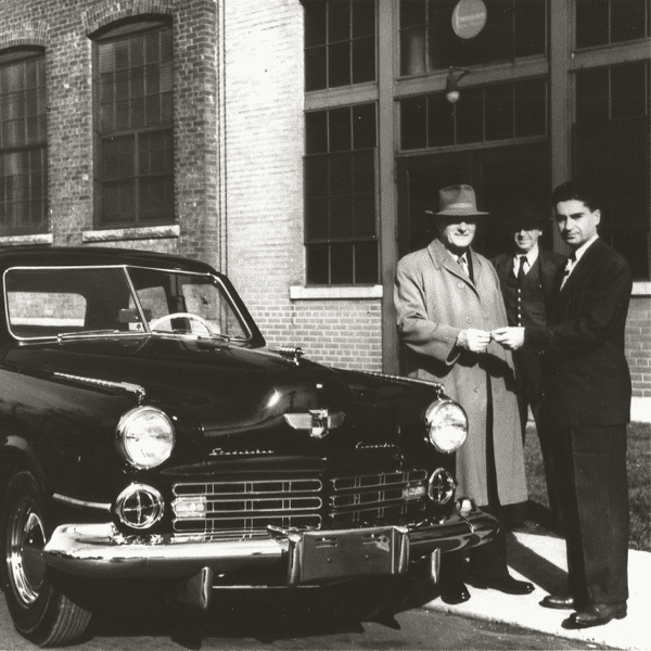 Black and white photograph depicting three men in suits standing next to a dark-coloured Studebaker car in front of a brick building. The man on the left is presumably handing a key to the man on the right. The other man is behind them.
