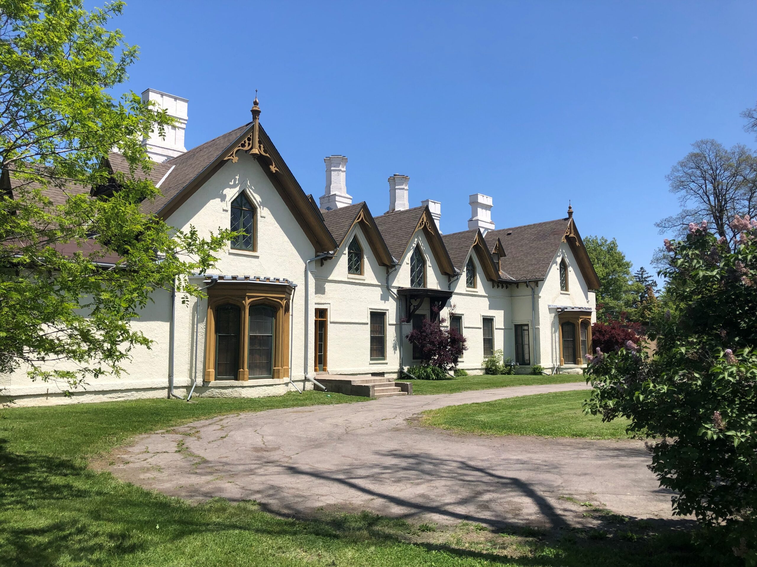 A photo of a white stucco manor house with gothic details. A curved driveway swoops past in the foreground. There are trees on either side of the house.