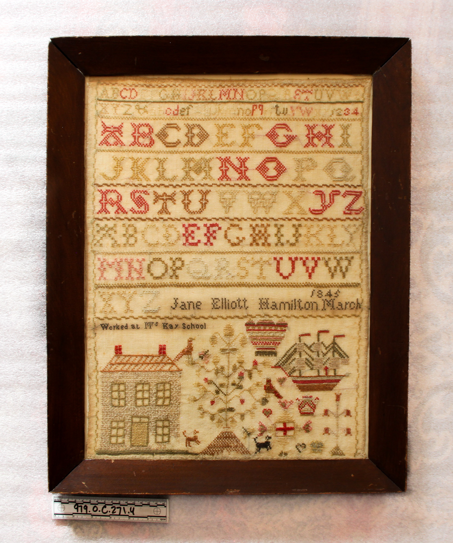 Image of a framed embroidery sampler, with the alphabet prominently cross stitched on the top half and images of a house, boat, and nature cross stitched at the bottom. A signature is cross stitched in the middle reading Jane Elliott Hamilton March 1845. A number is written on a label below the frame reading 979.O.C.271.4