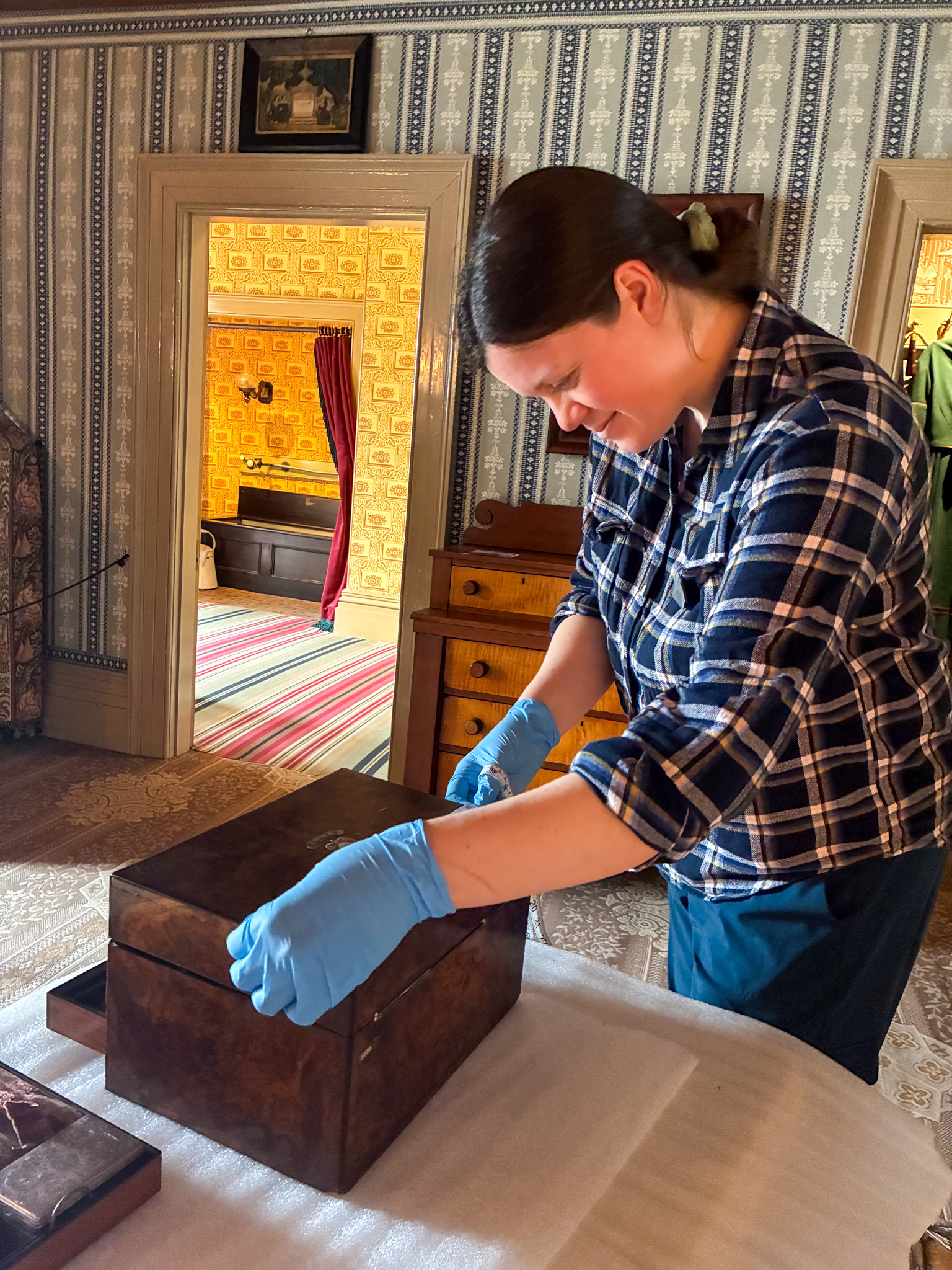 Image of staff member in a historic house room standing beside a table, measuring a wooden box that is sitting on top of foam with a tape measure.