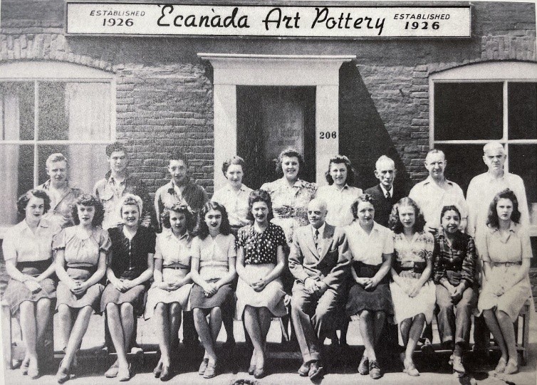 A black and white photograph of the employees of Ecanada Art Pottery, in front of their brick building workplace with the name of the company over the door. There are 2 rows of people, front row is seated. The people are wearing mearing clothing typical of mid-20th century craftsmen.