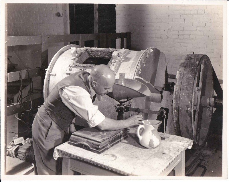 A black and white photo of a man (George Emery) leaning over a table next to a machine. He is measuring something. He is wearing a white shirt with the sleeves rolled and a vest.