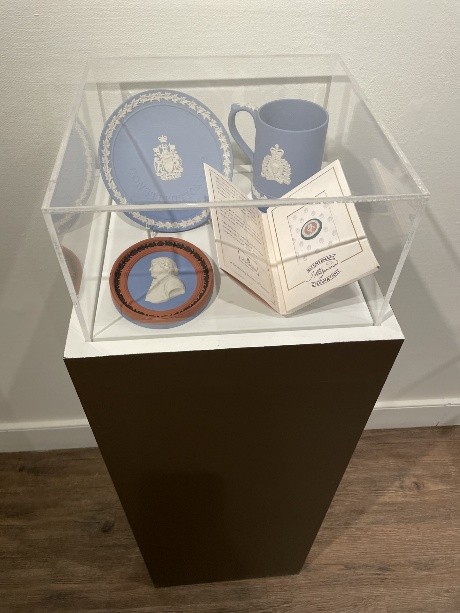 A display case features 3 classic Wedgwood pieces with the typical matte pale blue with withe details. There is also a small open booklet in the case.