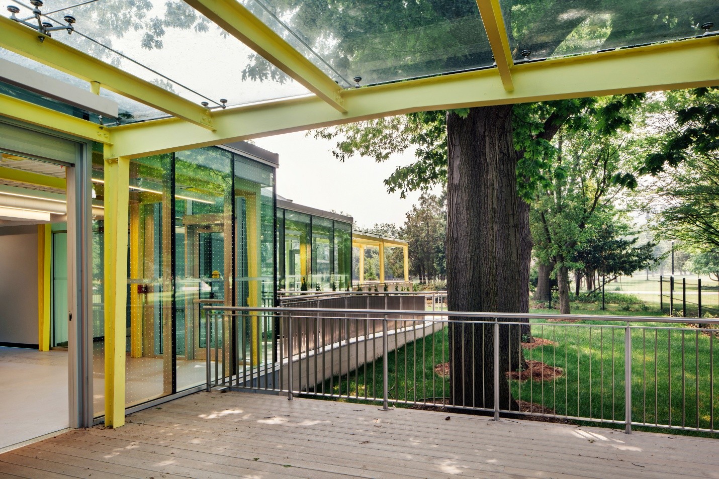 The outdoor covered space in the new backyard of the Children's Museum. There are yellow metal posts supporting a class roof over a wooden deck. The backyard has green grass and a large mature tree.
