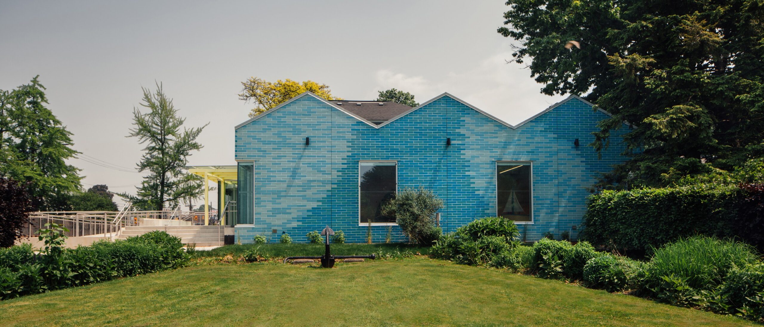 The new extension of the Children's Museum has several peaks and is made of shiny tiles in various shades of blue. It is surrounded by a garden and a tree.