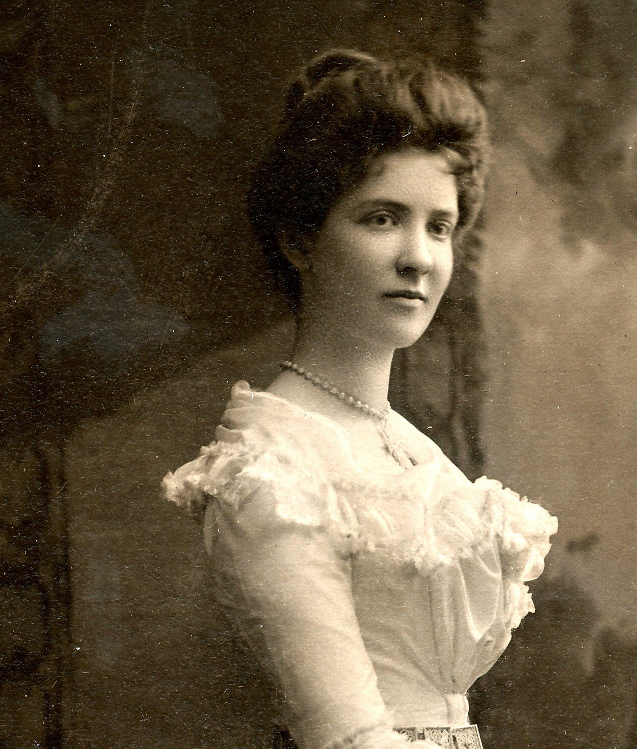 A black and white photo of a woman in the early 1900s. She has her hair up in a loose bun and is wearing a frilly white dress.