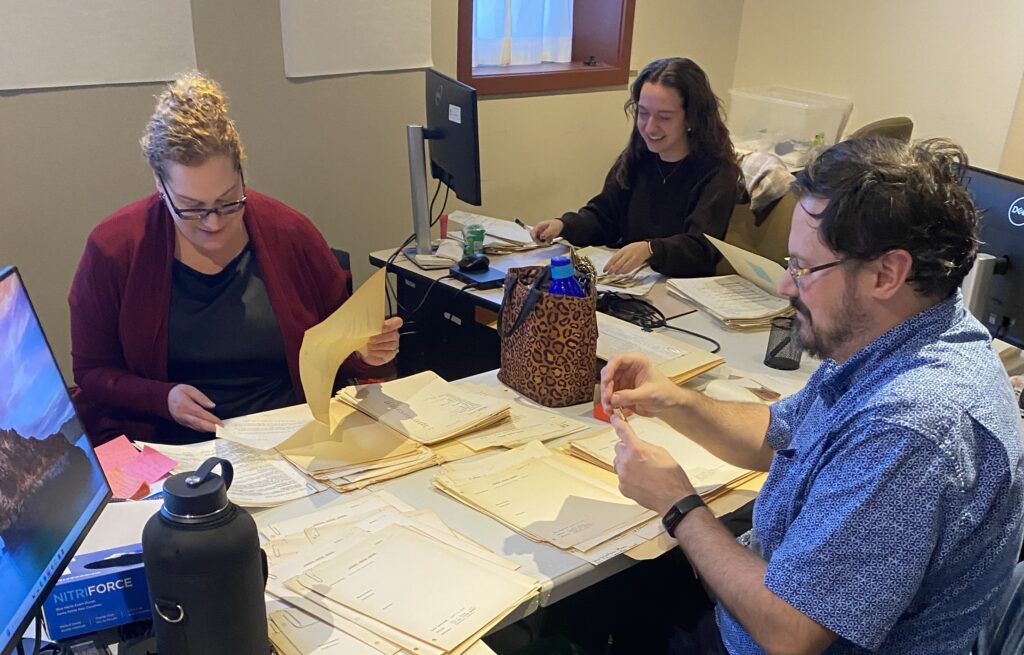 Three staff members sitting at their computer desks, sorting through piles of paperwork that sit on top of their workspaces.
