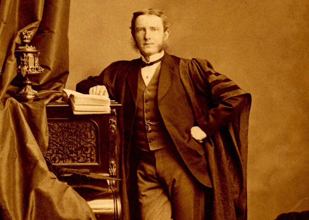 A sepia-tones black and white photo of a man wearing lawyers robes over a 3 piece suit. He is leaning on a n ornat piece of furniture which is draped by a curtain. It is in a photography studio.