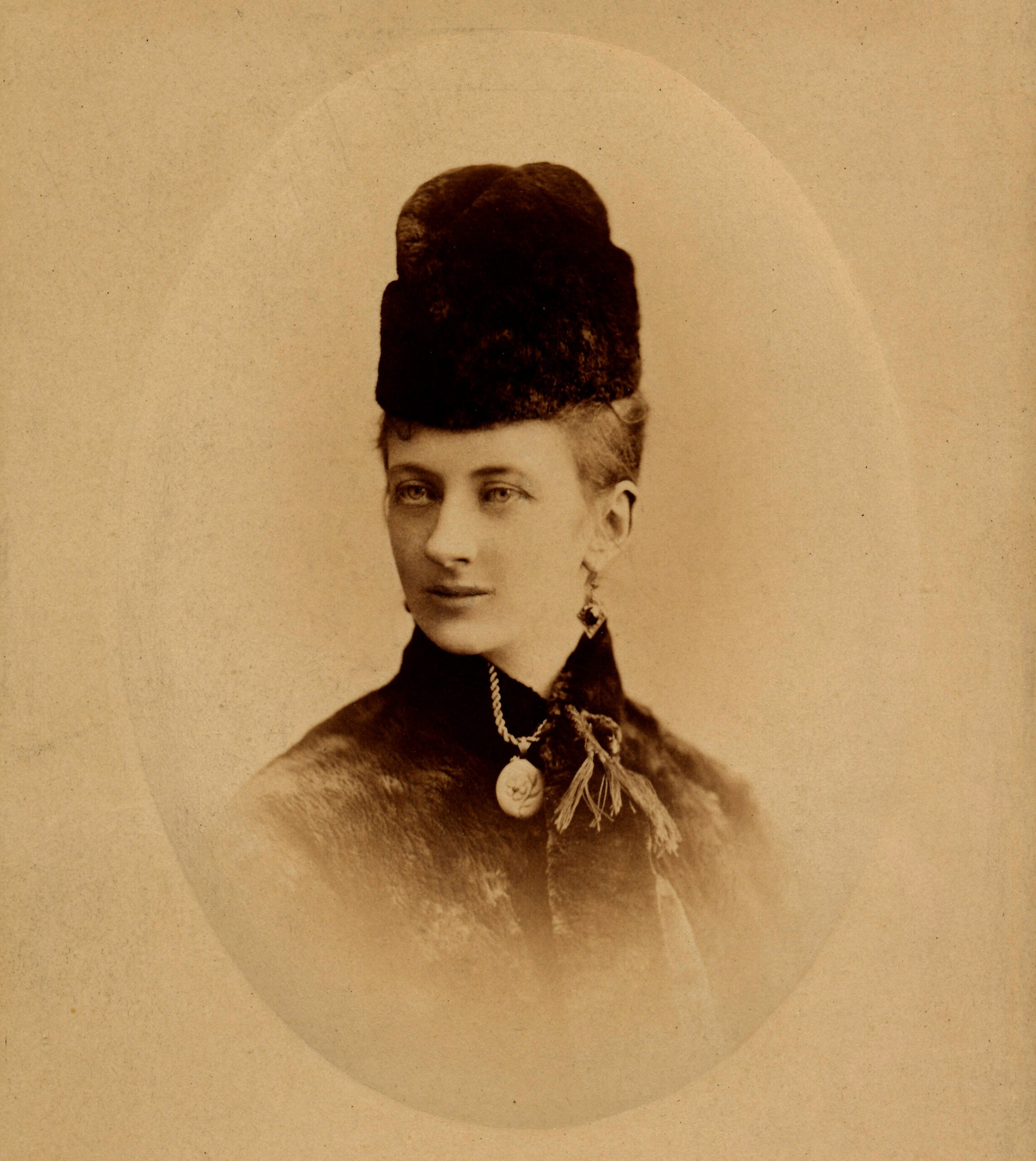 A sepia toned black and white photo of a woman. She is wearing a tall black hat and jacket.