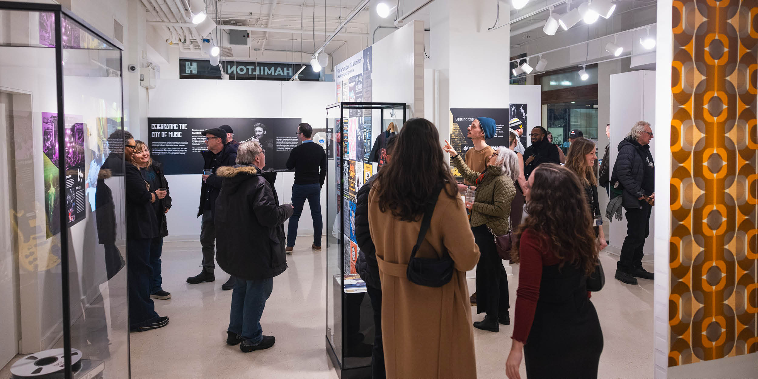 People are gathered in the exhibit. The photo shows over 10 people in small groups scattered around the space. There is a display case in the foreground on one side and orange and brown 1960s wallpaper on the right.