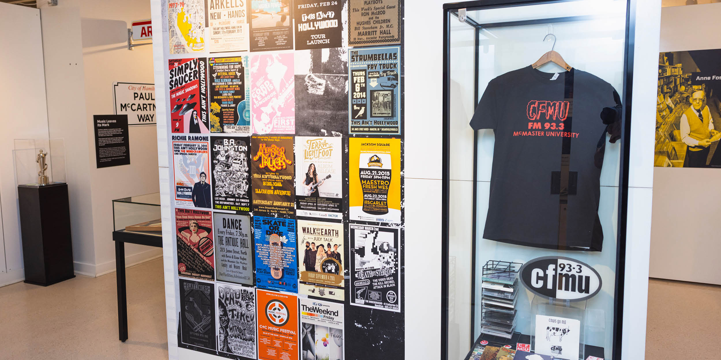 Part of the exhibit is pictured. A wall of music posters is next to a display case. In the display case is a black t shirt with CHMU's logo in red. There are more smaller artifacts below the t shirt.