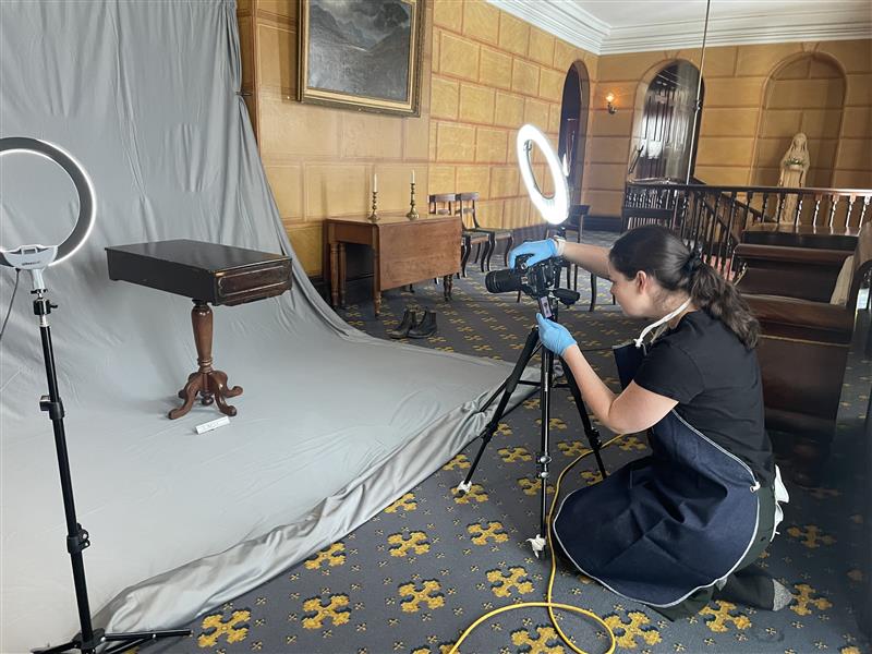 Image of a staff member crouching down behind a tripod and camera, photographing an antique table on a large grey backdrop surrounded by ring lights.