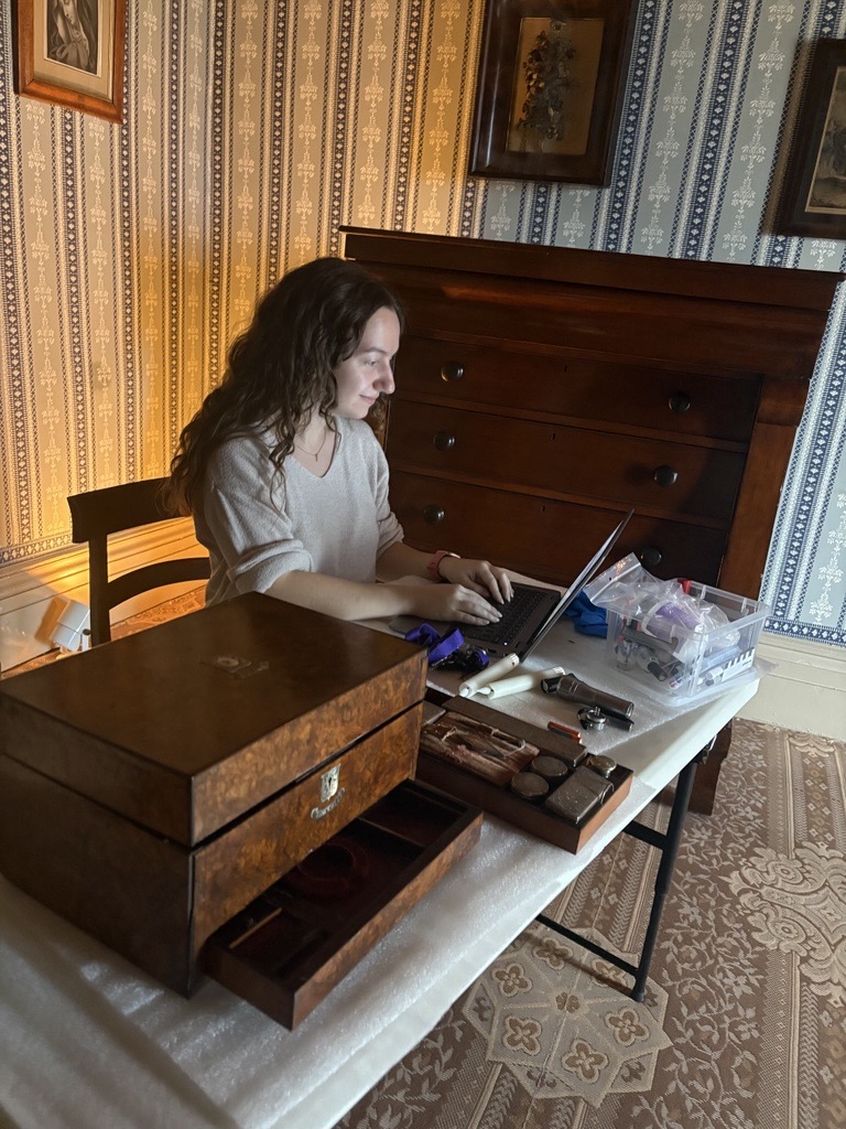Image of staff member in a historic house room sitting at a table, looking at a computer with a wooden artifact box beside her on the surface of the table.