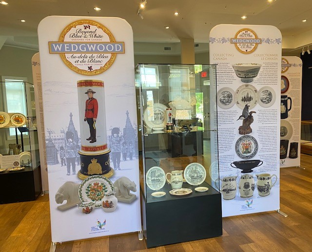 Two banners flank a display case. The banners display images of the artifacts in the exhibit, including pottery with Canadian iconography like a RCMP officer. The glass display case holds several plates.