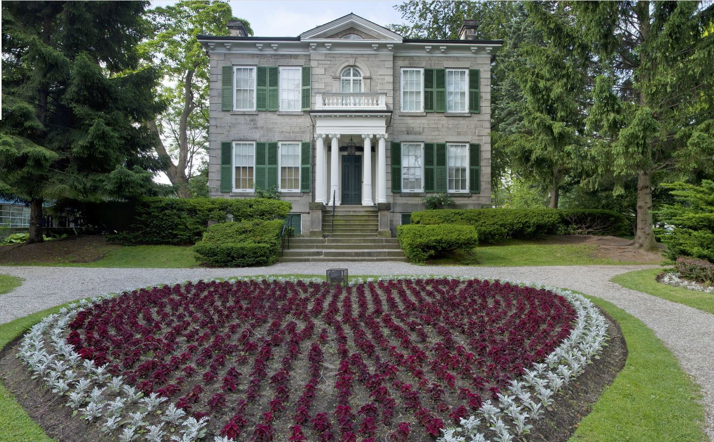 A modern photo of Whitehern. It is a grey stone house with green shutters and white columns in front of a black front door. In front of the house is a heart shaped garden bed with red flowers and a border of white flowers.