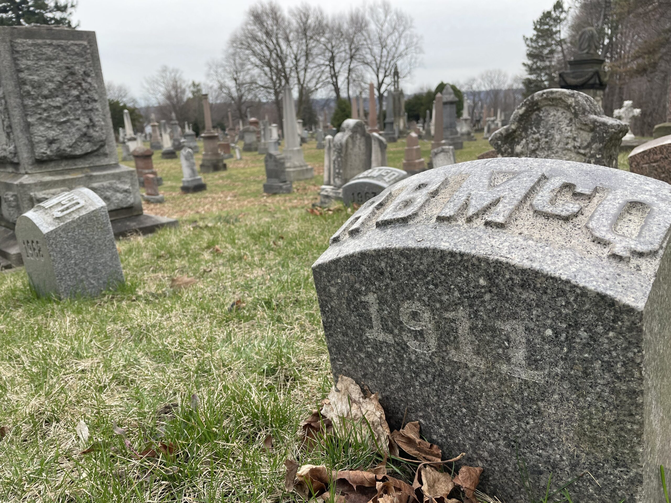 The small, round, grey grave marker of Thomas Baker McQuesten is in the foreground with more of Hamilton Cemetery in the backgroud.