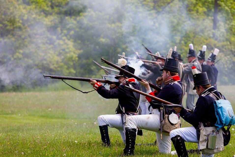 A group of male war of 1812 re-enacters in American uniforms fire muskets in a field, smoke and trees are behind them.