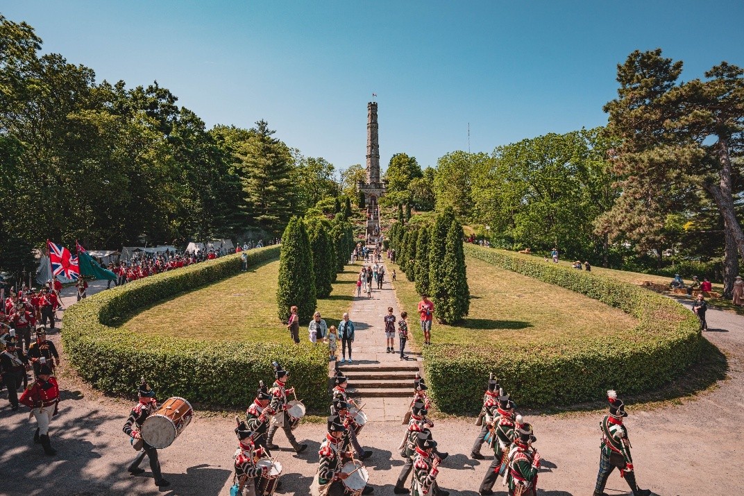 A parage of re-enactors in 1812 uniform march around a hedged lawn that leads up to the monument.