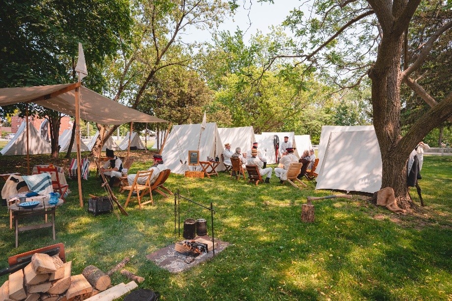 Wedge shaped white tents are arranged in rows in a re-enactment of a 1812 military camp. A campfire and wood are in the foreground.