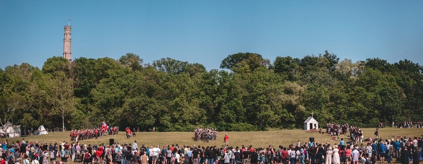 The Re-enactment of the Battle of Stoney Creek takes place between British and American re-enactors in the park next to Battlefield. The stone monument pokes up from behind trees in the background, and the audience stands in the foreground.