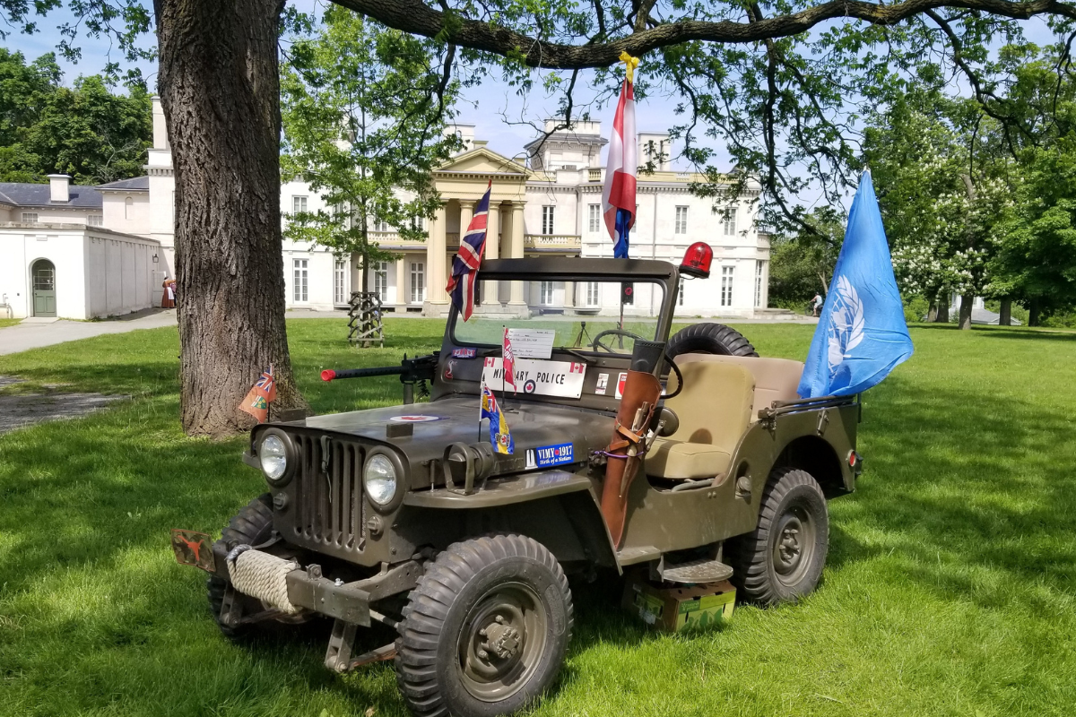 An open backed military vehicle with Canadian and United Nations flags is parked on the grass in front of Dundurn Castle under a tree.