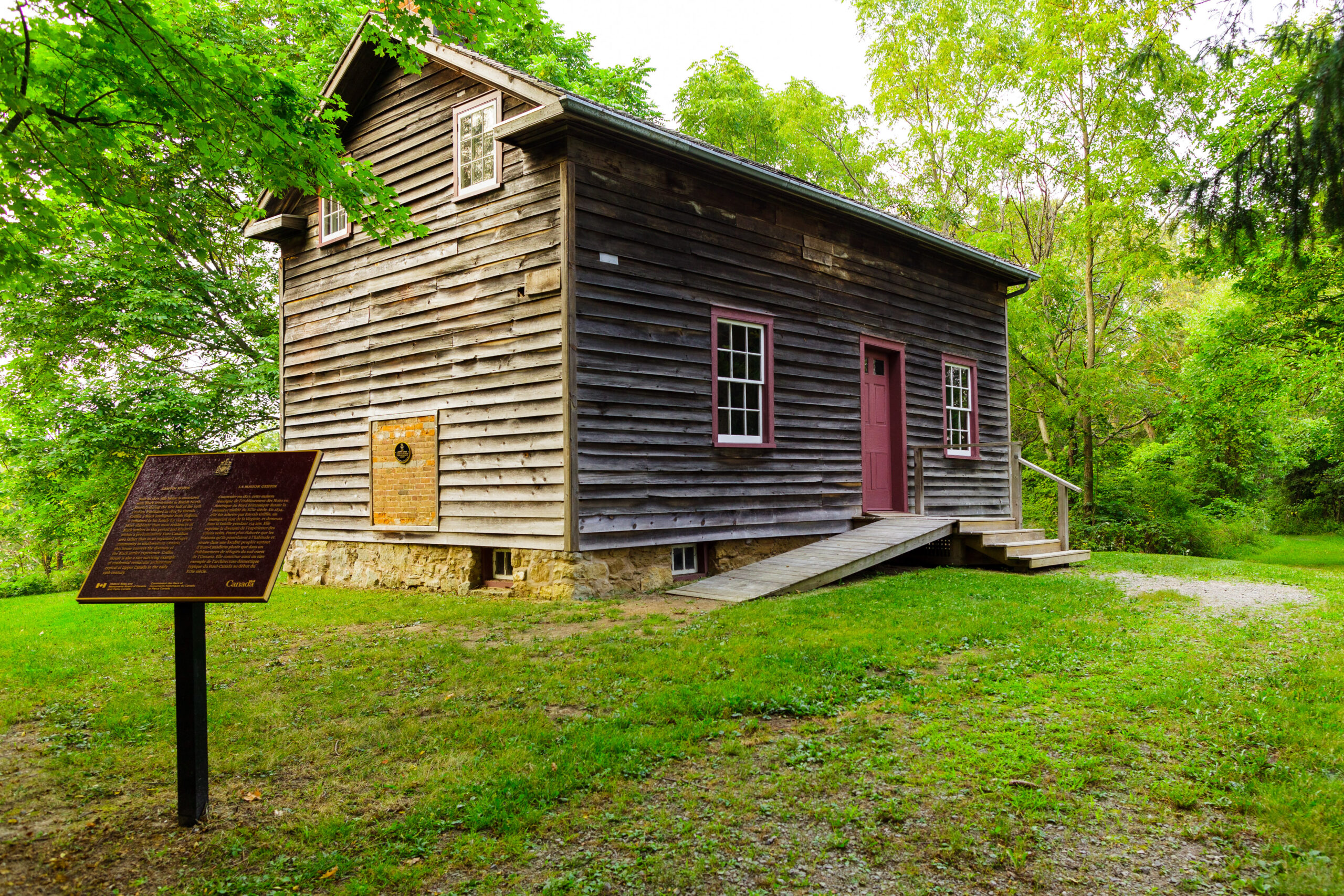 A one and a half story log cabin is surrounded by trees in a small clearing. It has dark wood siding and a red door. A commemorative plaque is in the foreground.
