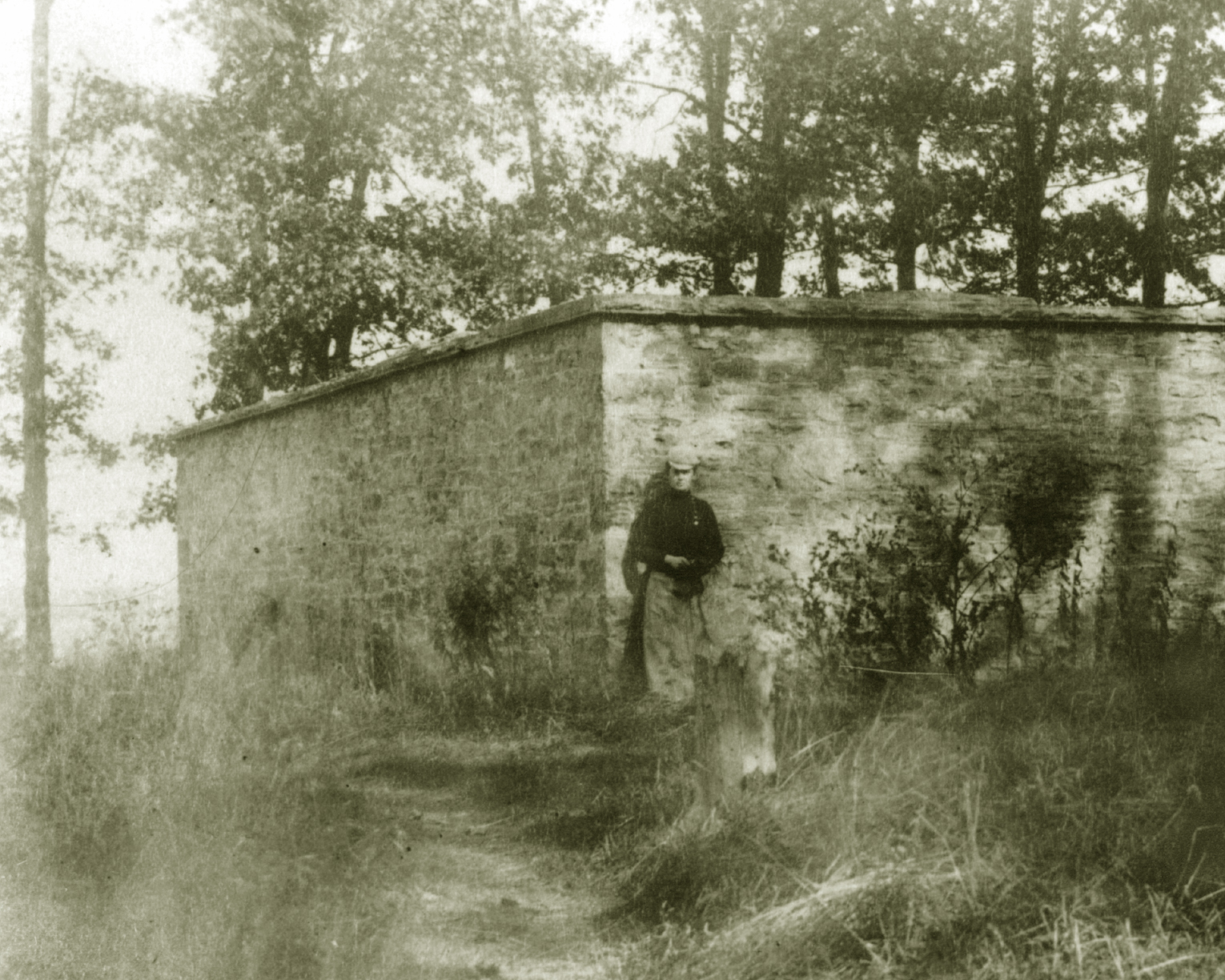 A black and white photo or a stone cube structure surrounded by trees. a man stands in front of the structure with long grass all around.