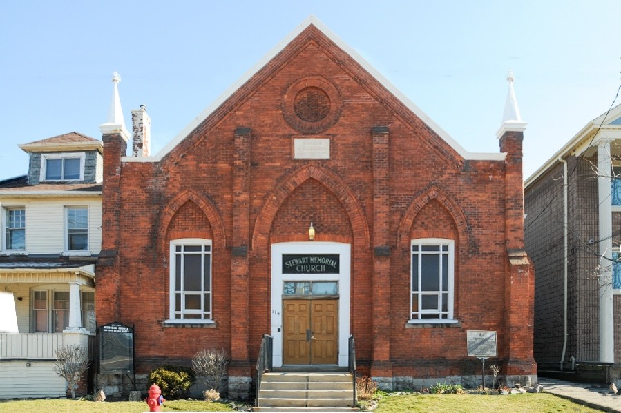 A red brick church. It is symmetrical with a white rectangular windows on either side of a light wood door. It does not have a church tower, it has a peaked roof.