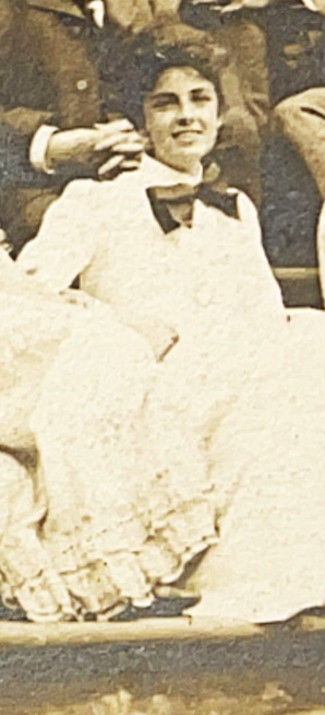A black and white photo of a woman wearing an Edwardian outfit of a white blouse with large black pussy-bow. She is semi-reclined on stone steps surrounded by other people that are cropped out. Her hair is in a Gibson-girl style bun.