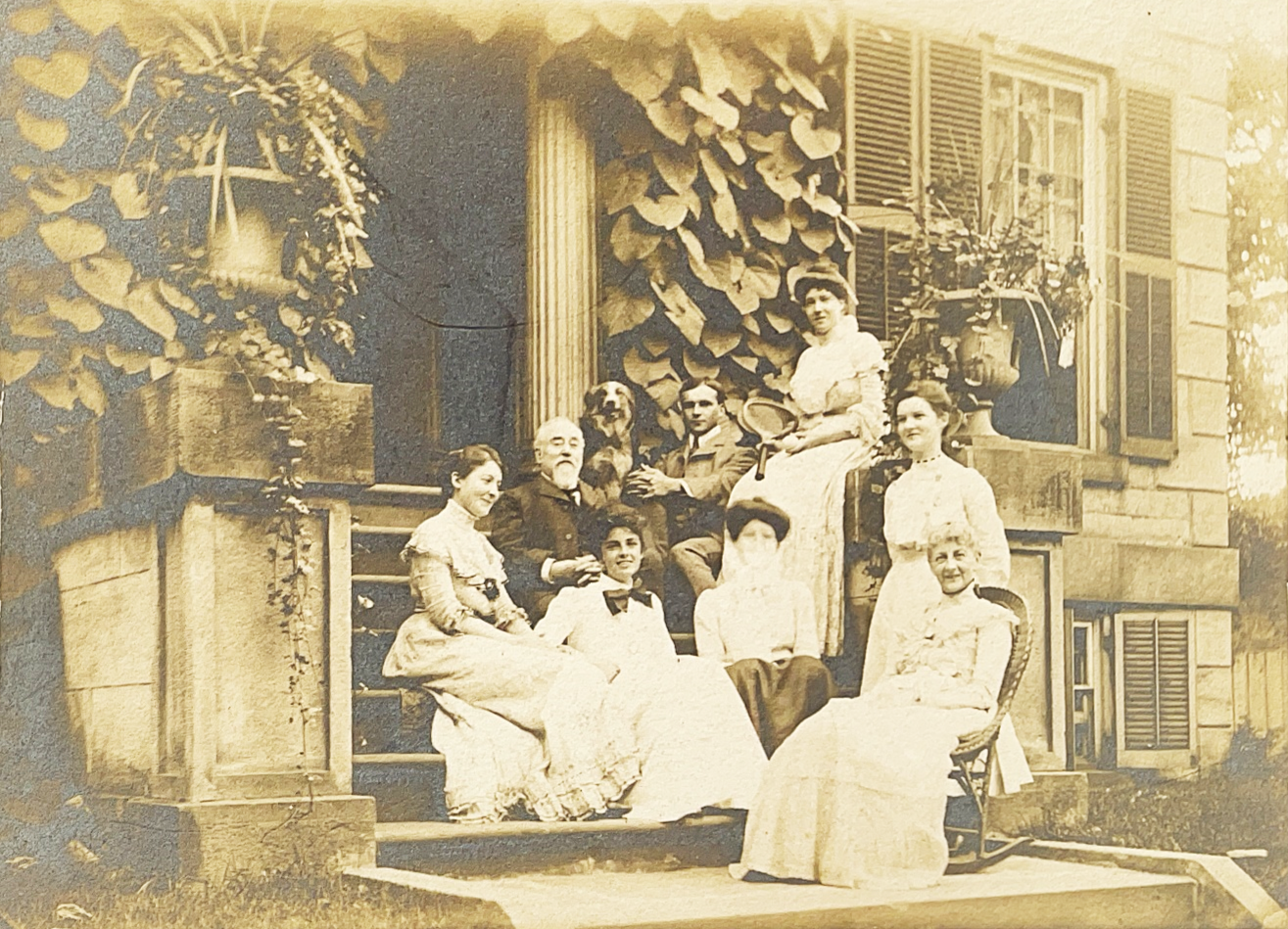 A black and white photo of group of people sit one and in front of the stone steps of Whitehern. It is the Edwardian era and most are wearing bright white.