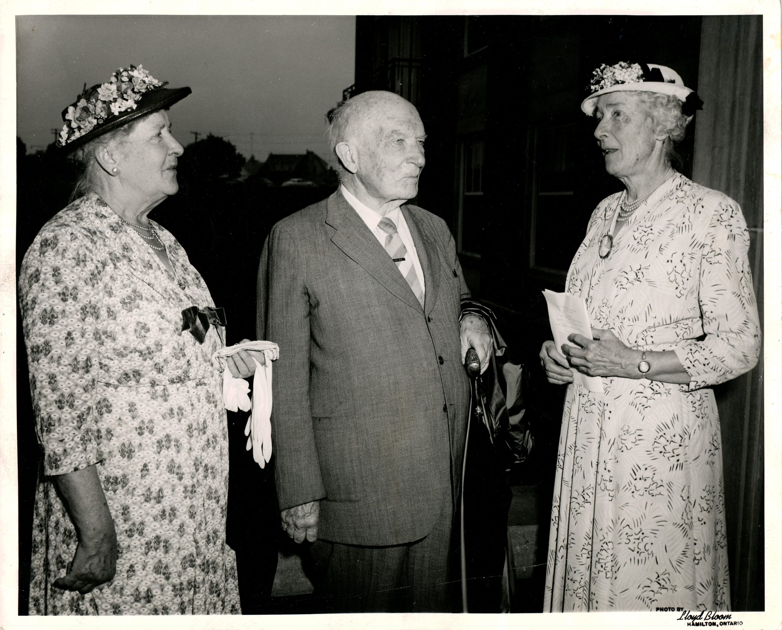 A black and white photo of 3 elederly people. The left is a woman in a floral dress and black sun hat with flowers. She is looking to the right. The centre is a man in a grey suit, also looking left. The right is a woman in a softer floral dress and white sun hat with flowers.