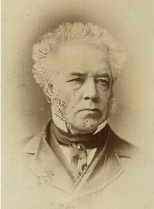 A black and white portrait with a sepia tone of a man. It is just his face and upper chest. He has white hair that sticks up and mutton chops. He looks over 50 years old and has a serious expression. He is dressed in mid 19th Century clothing.