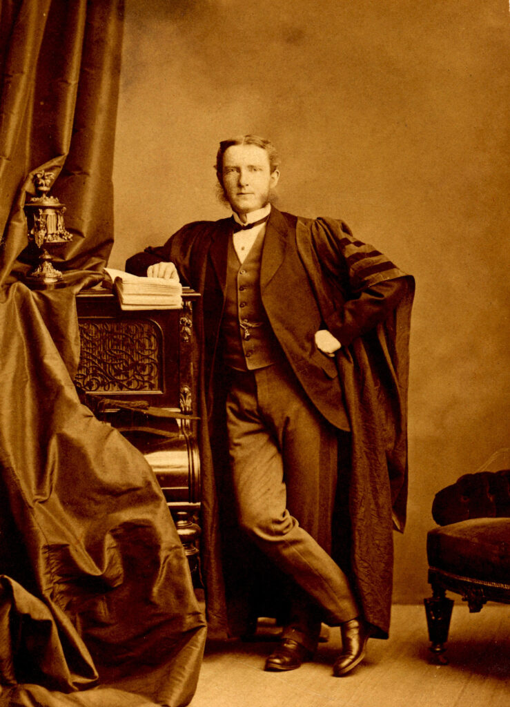 A sepia toned portrait of a man leaning on a piano that is draped with a curtain. It is taken in a photography studio, he is dressed in lawyer robes from the late 1800s.