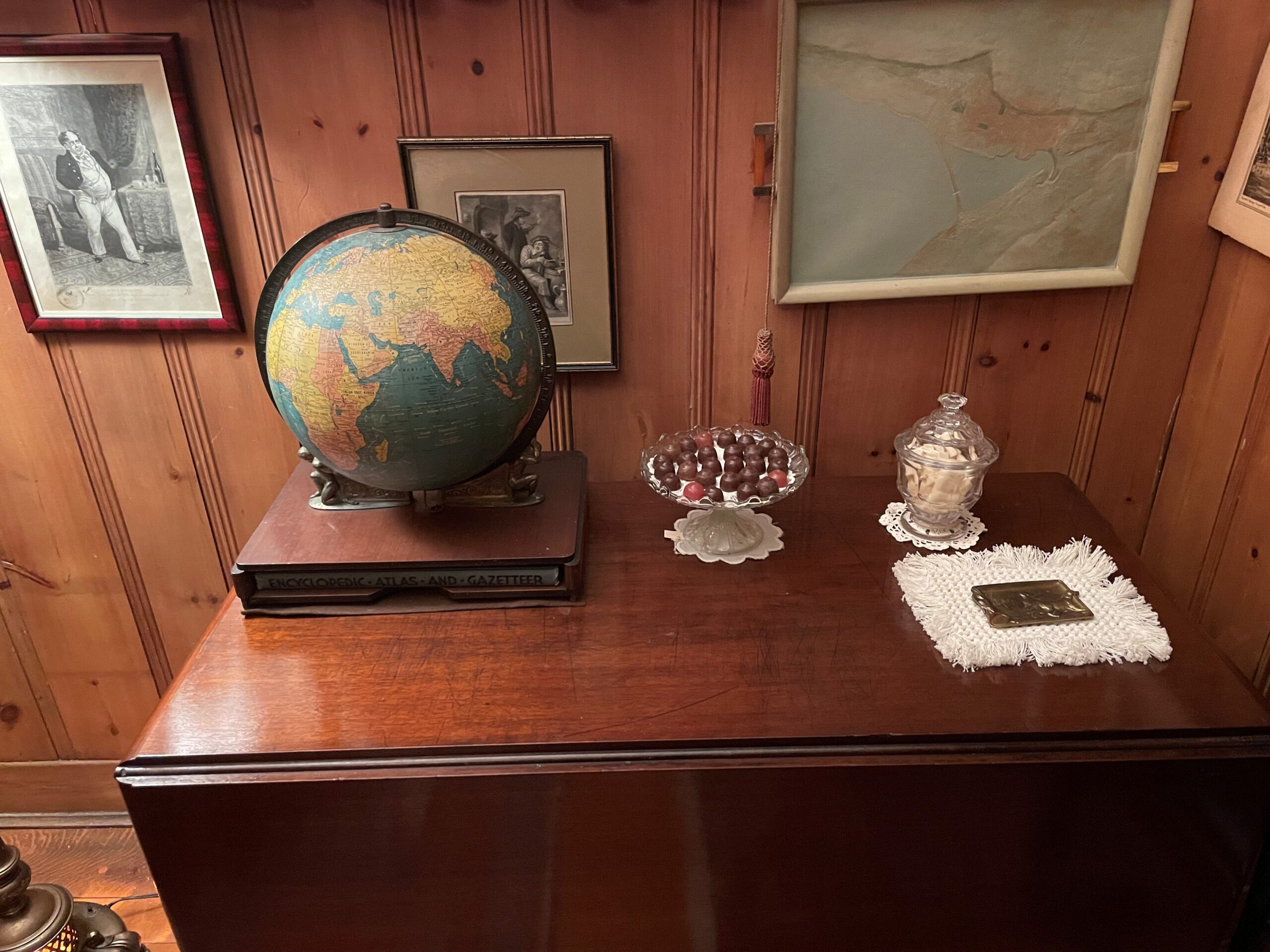 A photo of a tablescape. The walls are wood panelling with three framed photos. On the wood table is a vintage globe on a boxed encyclopedia, a crystal candy dish, a lidded crystal candy bowl and an ashtray. All are sitting on doilies