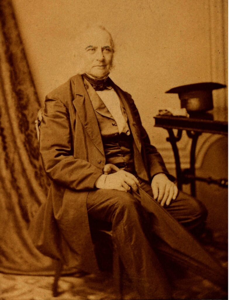 A sepia toned black and white image of a seated older man. He is in a photography studio with a curtain draped behind him to the left and a table with a hat to the right.