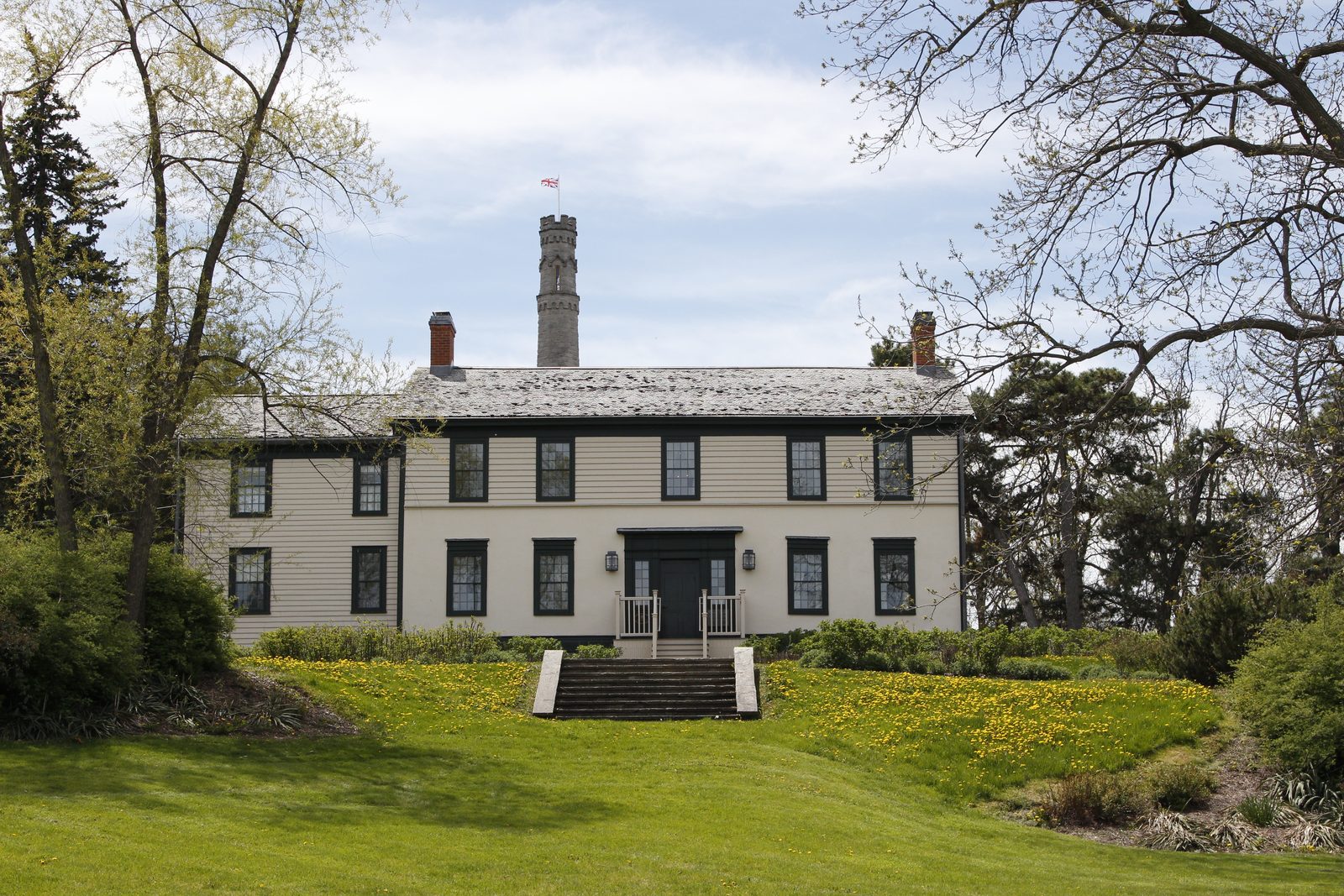 Battlefield House, a two-storey white clapboard house with dark green shutters, on a sloping lawn dotted with dandelions, with the stone Battlefield Monument tower rising behind it.