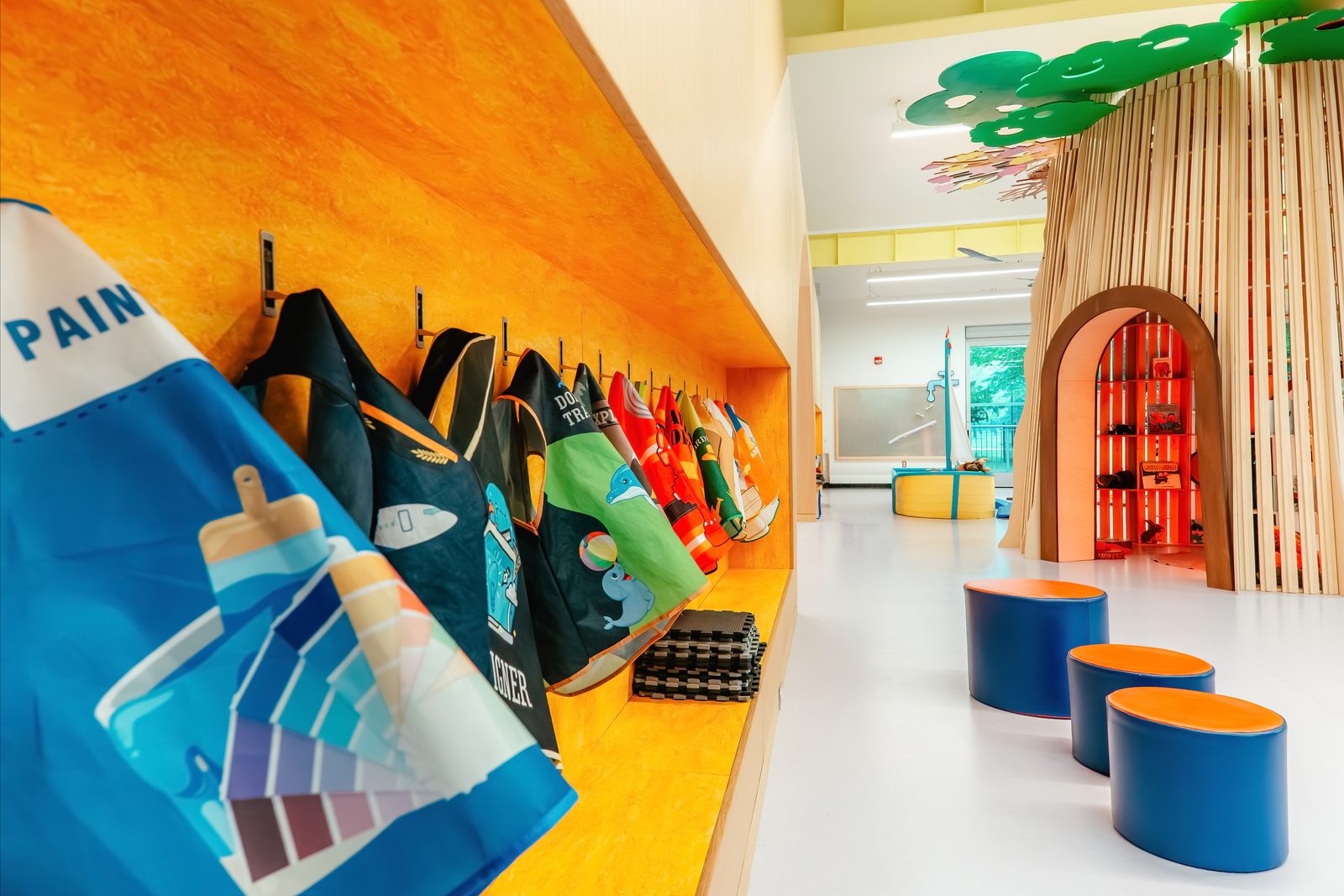 Colourful themed dress-up aprons hang on hooks along an orange wall inside the Hamilton Children's Museum, with a wooden treehouse play structure and blue stools in the background
