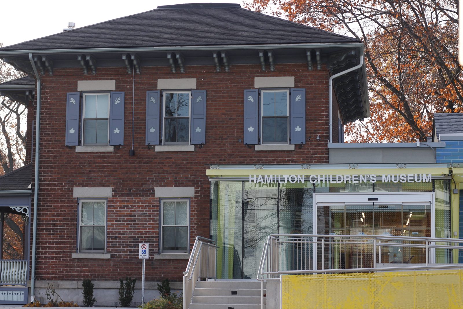 The Hamilton Children's Museum, a red-brick 1875 farmhouse with blue-grey shutters, and its glass entrance addition lettered Hamilton Children's Museum, with orange fall foliage behind.