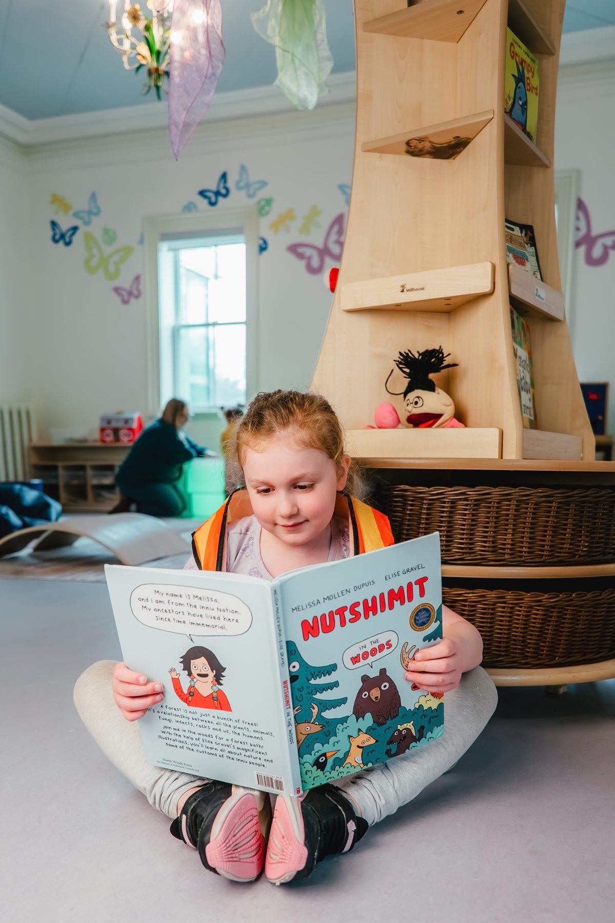 A young child in an orange safety vest sits cross-legged on the floor reading a picture book beside a wooden book tower in the Hamilton Children's Museum reading nook, with butterfly wall decals behind