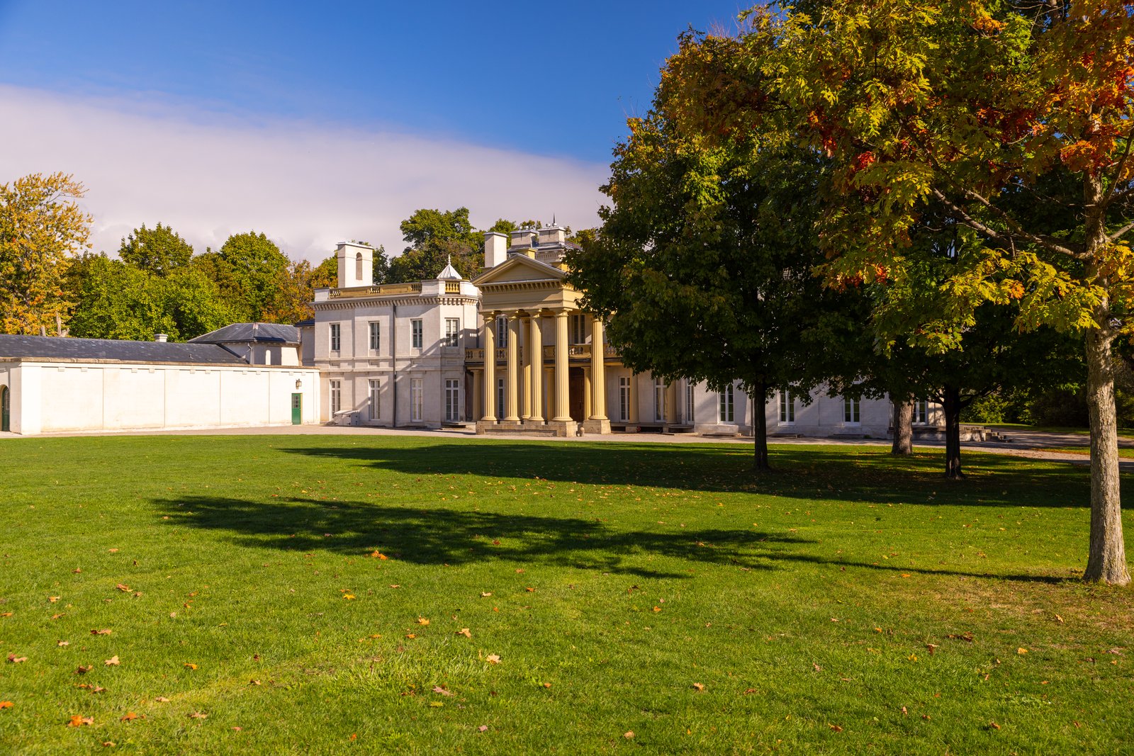 Dundurn Castle, a white Regency villa with a columned portico, across its wide front lawn on a sunny fall day, with a large tree turning orange in the foreground.
