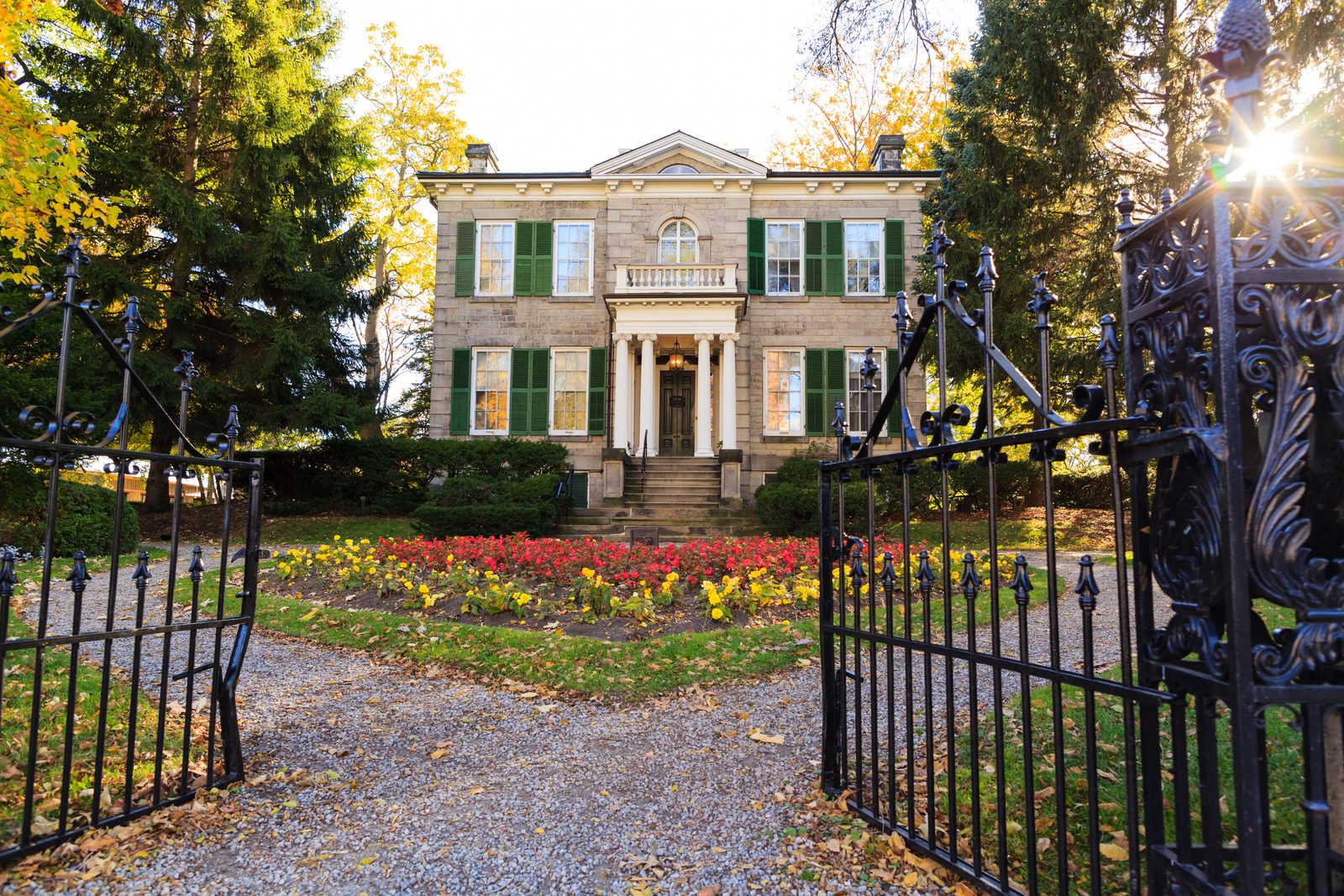 Whitehern, a two-storey stone house with green shutters, seen through its open iron gates with red and yellow flower beds and fall foliage.