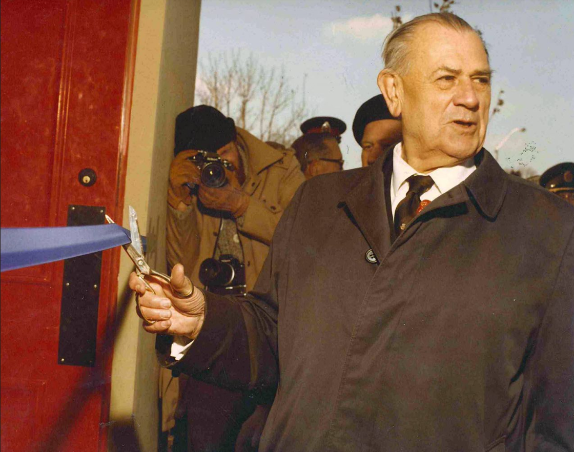 A colour photograph of a man (John Weir Foote) cutting a blue ribbon across a red door. He wears a shirt and tie and a dark grey coat.